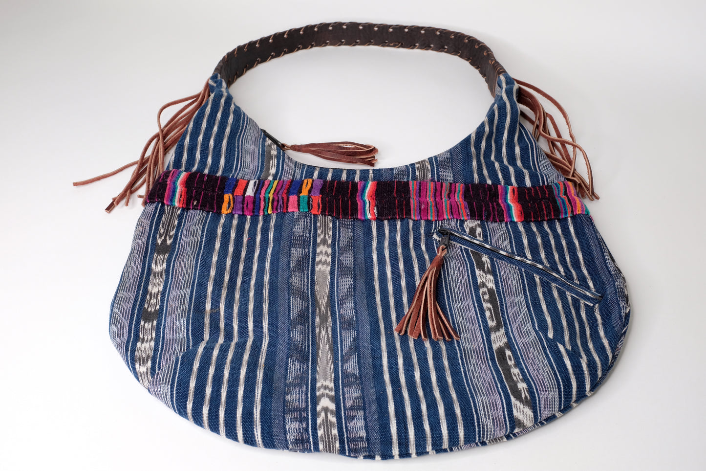 Blue handbag made with recycled Guatemala textiles, embroidery and leather fringe. Laying on a table.